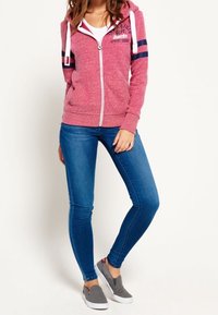 Donna con felpa rosa a zip con strisce, jeans skinny blu e scarpe slip-on grigie, in piedi con una gamba leggermente piegata.