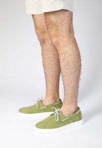 Grüne Wildlederschuhe mit weißen Gummisohlen, die eine runde Spitze, kontrastierende Nähte und dekorative Schnürsenkelösen an den Seiten aufweisen.