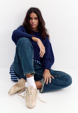 Jonge vrouw met lang donker haar die met gekruiste benen zit, draagt een marineblauwe trui, blauwe spijkerbroek, witte sokken en beige sneakers met losgemaakte veters.