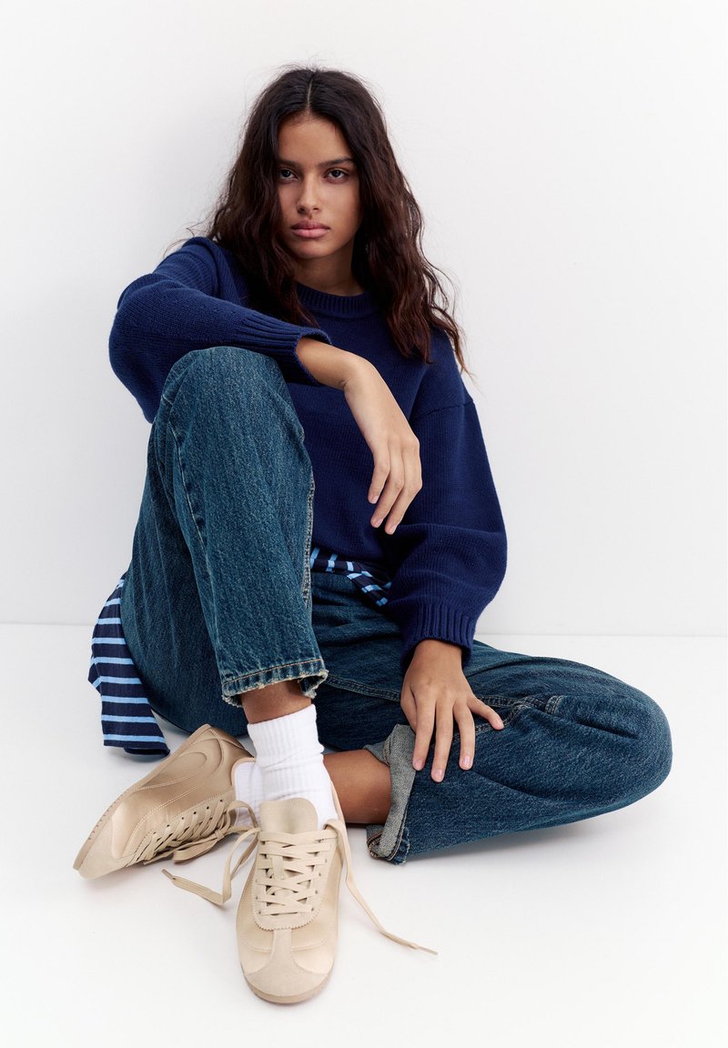 Jeune femme aux longs cheveux foncés assise en tailleur, portant un pull bleu marine, un jean bleu, des chaussettes blanches et des baskets beiges aux lacets défaits.
