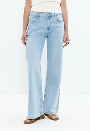 Femme portant un jean taille haute à jambes larges bleu clair, un haut court côtelé blanc, et des sandales marron à bouts ouverts, debout sur un sol blanc.