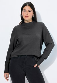 Frau mit langen dunklen Haaren, die hoop earrings trägt, einen dunkelgrauen Langarmpullover und schwarze Hosen anhat, steht mit einer Hand in der Tasche vor einer weißen Wand.