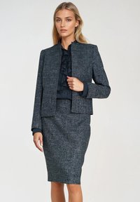 Kobieta w ciemnoszarej, teksturowanej garsonce z falbanistą czarną bluzką, pozująca z jedną ręką przy kurtce.