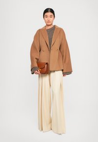 Blazer en laine marron à double boutonnage avec des manches oversize, porté sur un pull marron, associé à un pantalon large crème et un sac bandoulière marron.