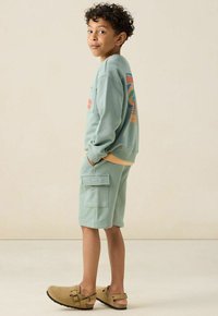 Sweatshirt vert menthe avec un design graphique, shorts assortis avec poches latérales, et sabots beiges avec une boucle décorative. Tenue décontractée dans l'ensemble.