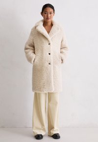 Crème-kleurige, getextureerde faux fur jas met een brede kraag, knopen aan de voorkant en zijzakken, gecombineerd met lichtgekleurde, gestreepte broek.