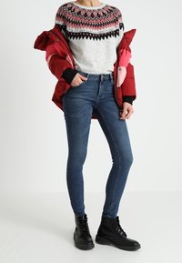 Punainen toppatakki, harmaa neulepaita, jossa vaaleanpunainen ja musta kuvio, siniset kapeat farkut, mustat nauhalliset saappaat, seisova asento valkoisella taustalla.