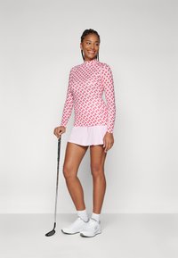 Pikemate golfipusa, millel on punane ja valge kootud muster, koos heleda roosa volangseelikuga ja valgete spordijalatsitega. Golfikepp käes.