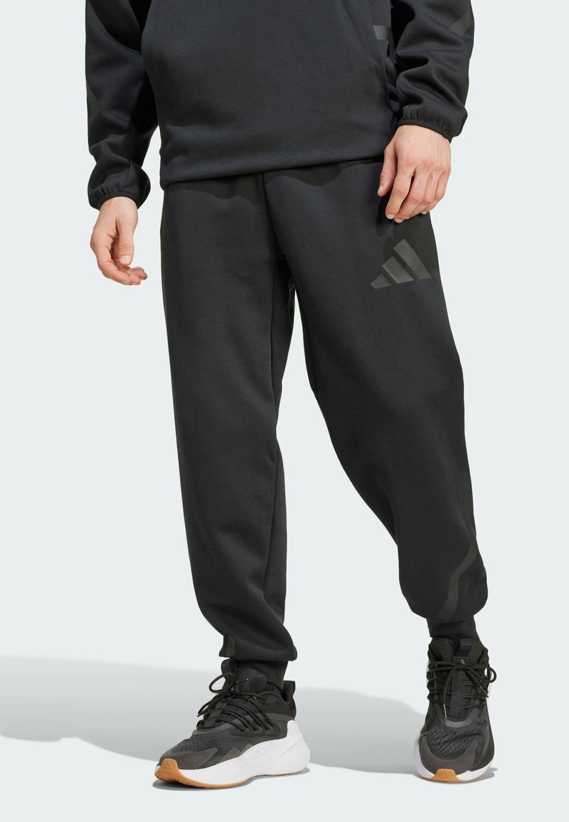 Black athletic pants made of soft fabric, featuring a tapered design, elastic cuffs, and subtle logo detail on the thigh; paired with black sneakers.