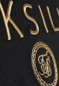 Tessuto nero con lettering dorato in rilievo e un logo ricamato circolare con dettagli intricati, che mette in mostra una superficie texturizzata.