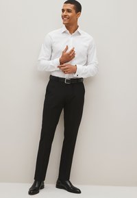 Witte blouse met lange mouwen en een neklap, gecombineerd met zwarte broek en zwarte klassieke schoenen. De blouse heeft strakke manchetten.