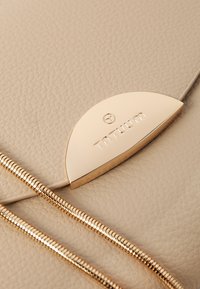 Beige lædertaske med et karakteristisk guldtonet halvcirkelformet detalje med "TATUUM" branding og to sammenflettede teksturerede stropper.