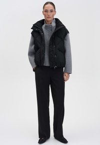 Schwarze gesteppte Daunenweste über einem grauen hochgeschlossenen Pullover, kombiniert mit hochgeschnittenen schwarzen Hosen und schwarzem Schuhwerk, vor einem schlichten Hintergrund.