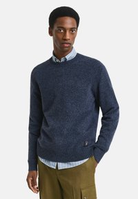 Marineblauer Strickpullover mit Rundhalsausschnitt, gerippten Bündchen und Saum. Über einem hellblauen, gestreiften Hemd getragen, das ein Etikett auf der linken Seite hat.
