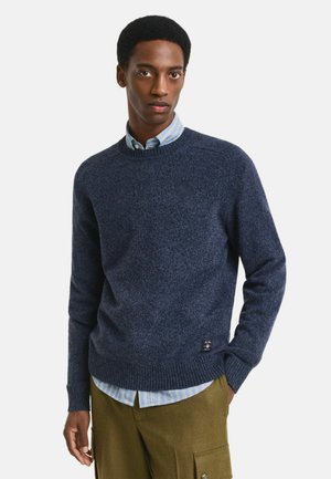 Marineblauwe gebreide trui met een ronde hals, geribde manchetten en zoom. Gelaagd over een lichtblauw gestreept shirt, met een label aan de linkerkant.