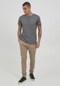 Szary, krótkim rękawem koszulka henley z trzema brązowymi guzikami, beżowe spodnie typu tapered i czarne sneakersy. Gładkie tło, stojąca postać mężczyzny.