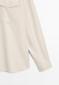 Chemise à manches longues de couleur crème avec une poche poitrine à boutons et un poignet à bouton unique, visible sur un fond blanc.