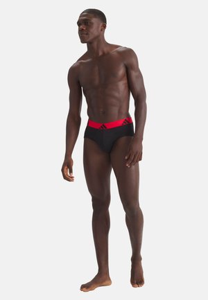 Uomo scuro in forma, in piedi a piedi nudi, che indossa slip neri con una cintura rossa con un logo nero, su uno sfondo chiaro e uniforme.