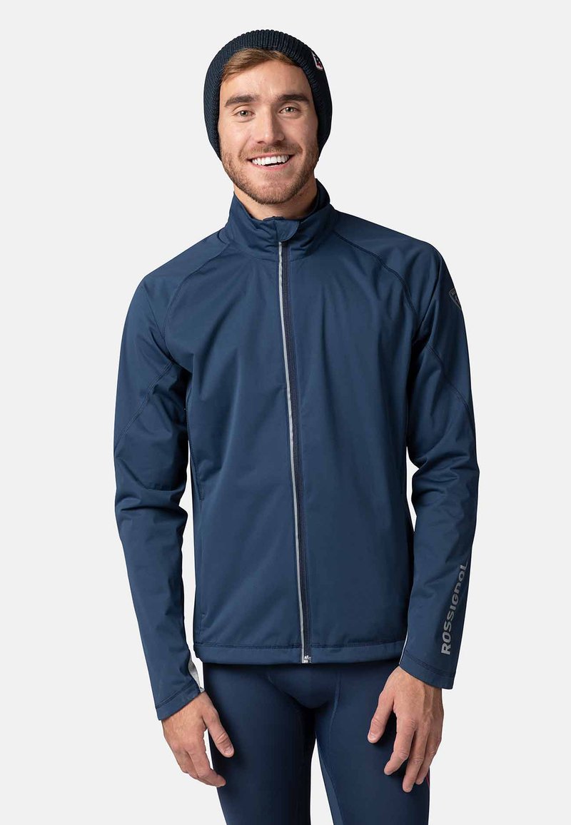 Marineblå lättviktjacka med hög krage, hel dragkedja framtill och kontrasterande grå detaljer. Har en logotyp på ärmen och en slät yta.