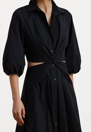 Žena v černých šatech na knoflíky s nafouknutými tříčtvrtečními rukávy a zkrouceným vykrojením v pase.