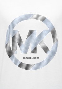 Camiseta blanca con un diseño de logo circular que presenta "MK" en tipografía a rayas negra y azul, con el texto "MICHAEL KORS" debajo.