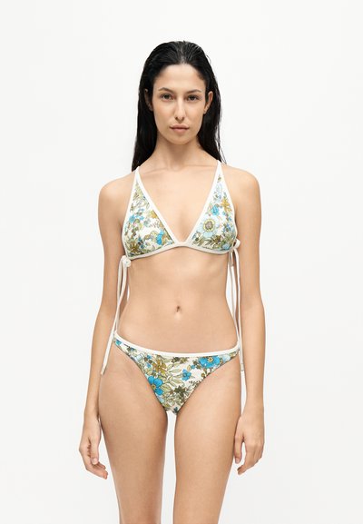 Femme aux longs cheveux foncés portant un bikini blanc à motif floral avec des fleurs bleues et vertes, debout devant un fond clair uni.