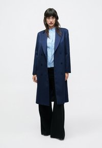 Tmavomodrý dvojradový kabát so širokými klopami, vyrobený z hladkej látky. Kombinovaný s svetlomodrou košeľou a čiernymi širokými nohavicami.