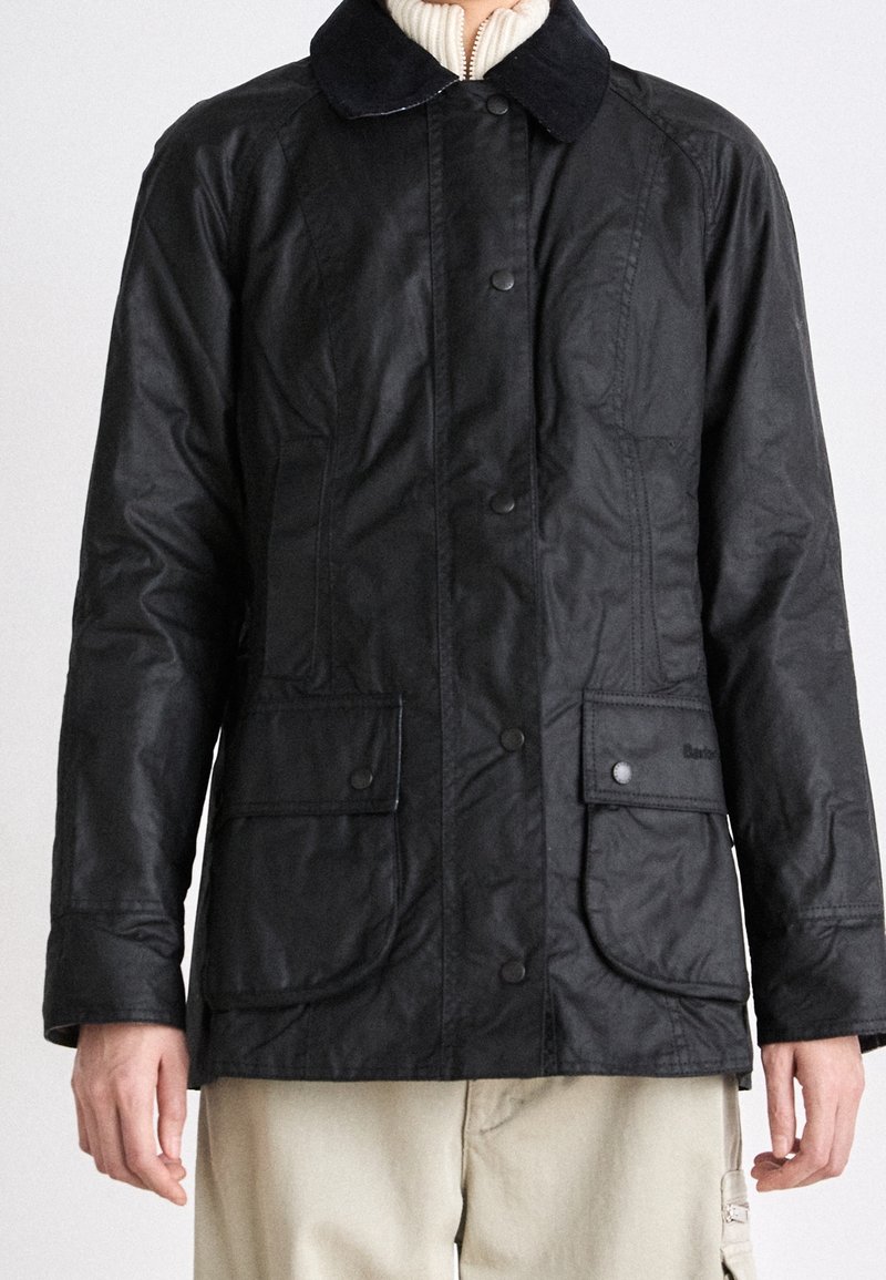 Personne portant une veste cirée noire avec un devant boutonné et deux grandes poches à rabat, associée à un pantalon beige et un pull en maille crème.