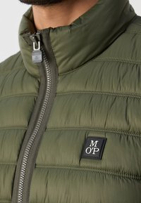 Närbild av en olivgrön vadderad jacka med frontdragkedja och en svart logotyp med texten "M O P" på bröstet.