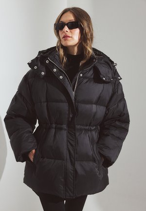 Sort pufferjakke med snøret talje, ydre lommer, aftagelig hætte og trykknaplukninger. Har en blød, blank tekstur.