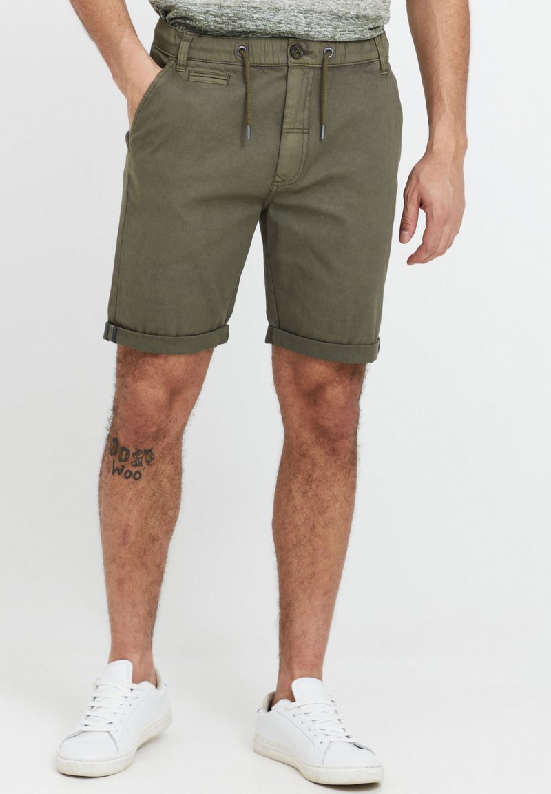 Shorts en coton vert olive avec des revers, poches avant et taille à cordon réglable, portés avec des baskets blanches.