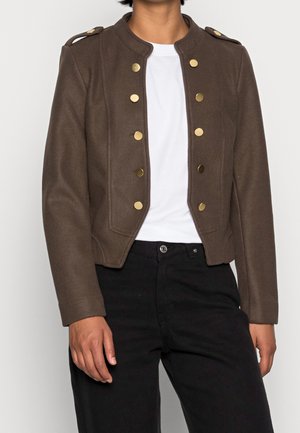 Osoba nosící tmavě hnědý krátký kabát se zlatými knoflíky přes bílou košili a černé kalhoty na jednobarevném pozadí.