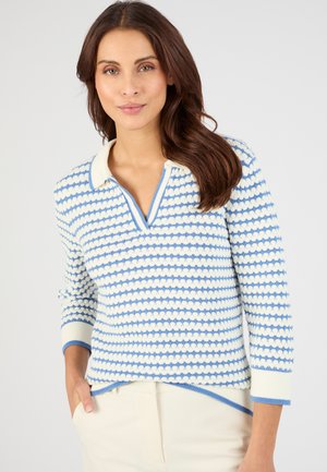 Femme aux longs cheveux bruns portant un pull en maille rayé blanc et bleu à col, avec un pantalon crème, posant avec une main dans la poche.