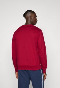 Mann med flettehår iført en enkel rød hettegenser og marineblå bukser med hvite side-striped, vendt bort fra en enkel bakgrunn.