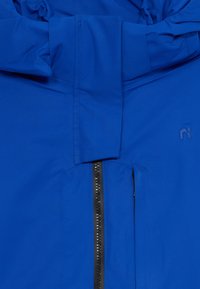 Chaqueta azul resistente al agua con cierre vertical, cuello alto y un discreto detalle de marca. Textura suave con costuras duraderas.