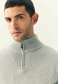 Joven con pelo corto y oscuro que lleva un suéter de cremallera de canalé gris claro con un cuello alto, sobre un fondo liso.