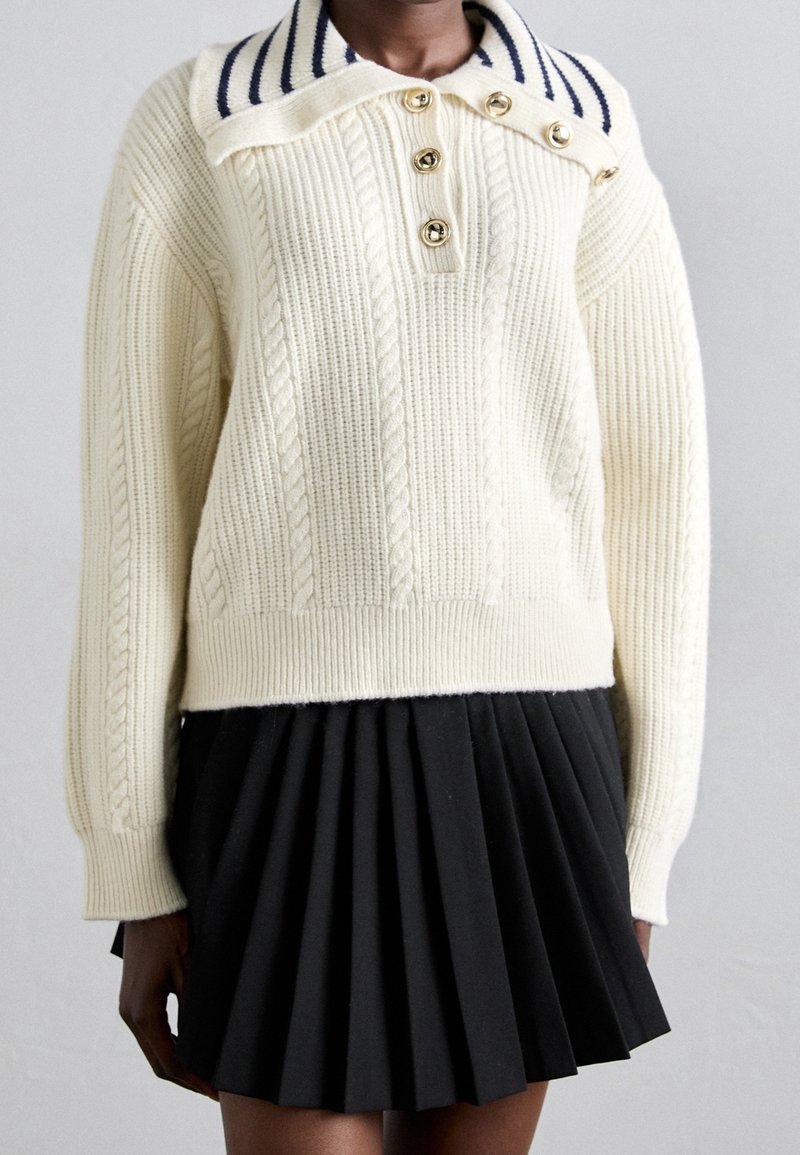 Cremefarbener Pullover mit Zopfmuster und einem kragen mit marineblauen Streifen sowie vier goldenen Knöpfen; kombiniert mit einem plissierten schwarzen Rock. Weiche Textur, lockere Passform.