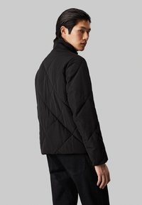 Chaqueta negra acolchada con cuello alto, diseño sin costuras y un acabado texturizado. Modelo mostrado de espaldas con jeans oscuros.