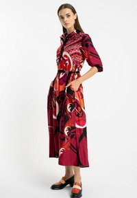 Kobieta stoi w kolorowej midi sukience z długim rękawem, ozdobionej odważnym abstrakcyjnym wzorem w czerwieni, pomarańczu i czerni, oraz w czarnych i pomarańczowych butach na niskim obcasie.