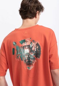T-shirt overdimensionato arancione in cotone, con una grande stampa grafica sul retro caratterizzata da colori molteplici e illustrazioni dettagliate.