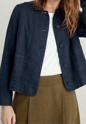 Femme portant une veste bleu marine boutonnée par-dessus une chemise blanche et une jupe vert olive, avec de longs cheveux ondulés couvrant partiellement son visage.