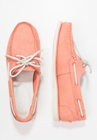 Korallmocka moccassinsläpare med vita snören och sålar, visade från ovan- och sidovy på en vit bakgrund.