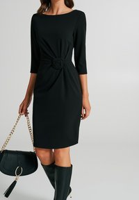 Robe noire avec des manches trois-quarts, détail de taille torsadé ressemblant à une rose, assortie avec un sac à main noir et des bottes montantes.