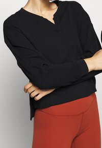 Schwarzes langärmeliges Rippenoberteil mit einem Henley-Ausschnitt, das einen lässigen Schnitt und kontrastierende Bündchen hat. Getragen mit rötlich-braunen Leggings.