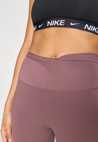 Μαύρο αθλητικό σουτιέν Nike με λευκή λωρίδα λογότυπου, συνδυασμένο με ψηλόμεσα κολάν σε μωβ, με απαλή υφή και αόρ ατο σχέδιο.