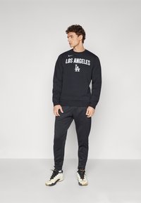 Melna sporta krekls ar baltu uzrakstu "LOS ANGELES", apvienots ar melniem sporta biksēm un bēšiem apaviem. Vienkārša dizaina, kokvilnas maisījuma materiāls.