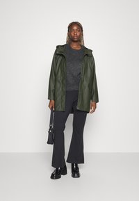 Veste imperméable verte avec capuche, associée à un pull gris foncé et un pantalon évasé noir. Le modèle tient un petit sac noir.