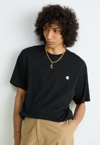 Black cotton t-shirt with a loose fit and short sleeves. Features a small white logo on the left chest. Paired with beige pants.