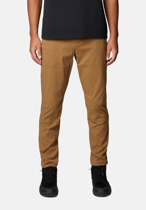 Mann trägt braune Slim-Fit-Hose und schwarze High-Top-Sneaker, steht mit locker hängenden Händen seitlich.