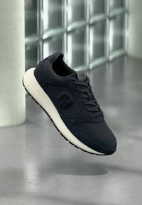 Σκούρο μπλε ναυτικό αθλητικό παπούτσι με συνδυασμό υλικών από διχτυωτό ύφασμα και σουέντ, λευκή μεσαία σόλα και υφή μαύρης καουτσούκ σόλας· διαθέτει λογότυπο στο πλάι.